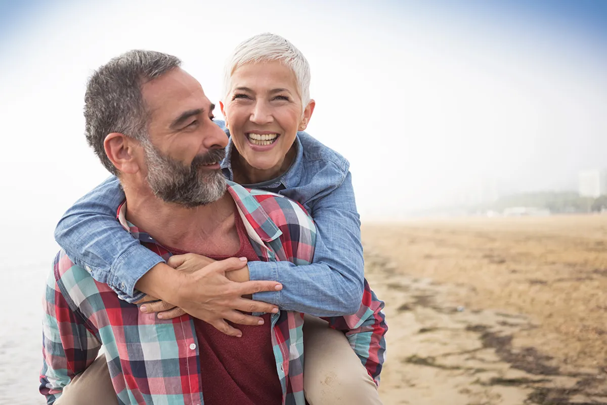 A middle aged man is giving a lady of a similar age a piggyback along the beach whilst they both smile