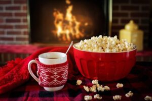 A mug with a red knitted cosy beside a bowl of popcorn in front of a fire