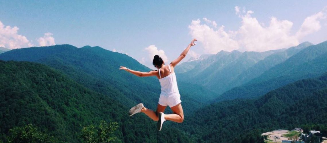 A lady jumping high in the air with her arms out wide above a green mountain valley